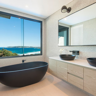 Diseño de cuarto de baño contemporáneo con lavabo sobreencimera, armarios con paneles lisos, puertas de armario de madera clara, bañera exenta, baldosas y/o azulejos grises y baldosas y/o azulejos en mosaico