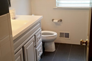 ワシントンD.C.にある高級な中くらいなトラディショナルスタイルのおしゃれなバスルーム (浴槽なし) (レイズドパネル扉のキャビネット、グレーのキャビネット、アルコーブ型シャワー、一体型トイレ	、白いタイル、磁器タイル、グレーの壁、セラミックタイルの床、一体型シンク、クオーツストーンの洗面台、グレーの床、開き戸のシャワー) の写真