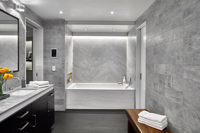Modern inredning av ett en-suite badrum, med släta luckor, skåp i mörkt trä, ett undermonterat badkar, en dusch i en alkov, en vägghängd toalettstol, grå kakel, stenkakel, grå väggar, mörkt trägolv, ett undermonterad handfat och marmorbänkskiva
