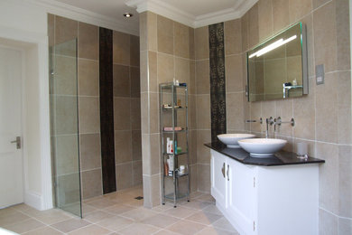 Expansive contemporary ensuite bathroom in Cornwall with a vessel sink, freestanding cabinets, white cabinets, granite worktops, a freestanding bath, an alcove shower, a wall mounted toilet, beige tiles, stone tiles, beige walls and travertine flooring.