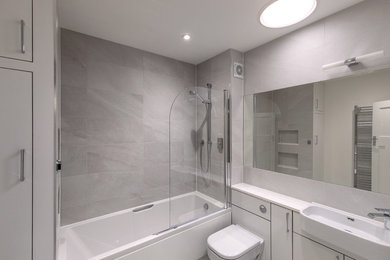 Photo of a medium sized contemporary family bathroom in London with flat-panel cabinets, grey cabinets, a built-in bath, a shower/bath combination, a wall mounted toilet, grey tiles, porcelain tiles, white walls, porcelain flooring, a built-in sink, solid surface worktops, grey floors and white worktops.