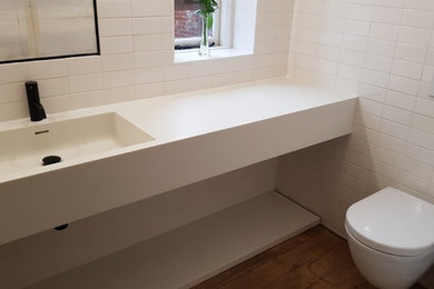 Small contemporary family bathroom in Gloucestershire with white cabinets, a wall mounted toilet, white tiles, ceramic tiles, medium hardwood flooring, an integrated sink, solid surface worktops, beige floors and white worktops.
