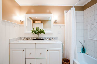Modelo de cuarto de baño de estilo americano con lavabo encastrado, armarios con paneles con relieve, puertas de armario blancas, encimera de mármol, combinación de ducha y bañera, paredes blancas, aseo y ducha y suelo de baldosas de porcelana