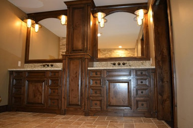 シカゴにある広いラスティックスタイルのおしゃれなマスターバスルーム (レイズドパネル扉のキャビネット、濃色木目調キャビネット、茶色い壁、トラバーチンの床、御影石の洗面台、茶色い床、ベージュのカウンター) の写真