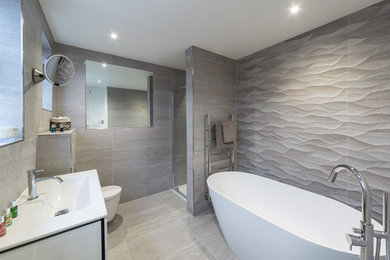 This is an example of a medium sized contemporary ensuite bathroom in Surrey with louvered cabinets, white cabinets, a freestanding bath, a corner shower, a wall mounted toilet, grey tiles, porcelain tiles, grey walls, porcelain flooring, a wall-mounted sink, solid surface worktops, grey floors, a hinged door and white worktops.