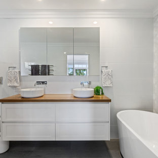 This is an example of a beach style bathroom in Central Coast with shaker cabinets, white cabinets, a freestanding tub, white tile, a vessel sink, wood benchtops, brown benchtops, a double vanity, a floating vanity, a one-piece toilet and grey floor.