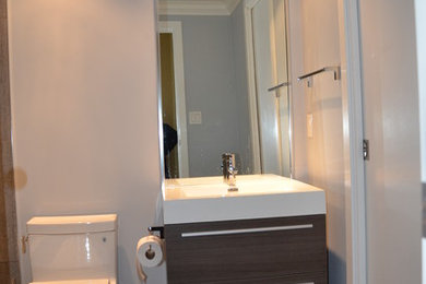 Klassisk inredning av ett litet badrum, med ett undermonterad handfat, släta luckor, skåp i mellenmörkt trä, bänkskiva i akrylsten, ett badkar i en alkov, en dusch/badkar-kombination, en toalettstol med hel cisternkåpa, brun kakel, porslinskakel, grå väggar och klinkergolv i porslin