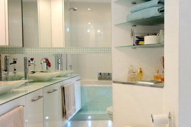 Small modern bathroom in London with a vessel sink, flat-panel cabinets, white cabinets, glass worktops, an alcove bath, green tiles, mosaic tiles, white walls and ceramic flooring.