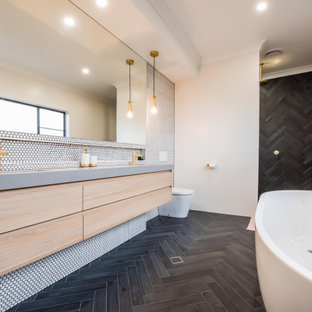 This is an example of a contemporary master bathroom in Sydney with engineered quartz benchtops, flat-panel cabinets, light wood cabinets, a freestanding tub, a one-piece toilet, white tile, mosaic tile, white walls, a wall-mount sink, black floor, an open shower and grey benchtops.