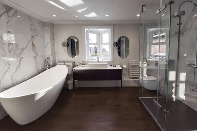 Photo of a medium sized contemporary ensuite bathroom with flat-panel cabinets, dark wood cabinets, a freestanding bath, a wall mounted toilet, beige tiles, marble tiles, beige walls, dark hardwood flooring, quartz worktops, a hinged door and white worktops.