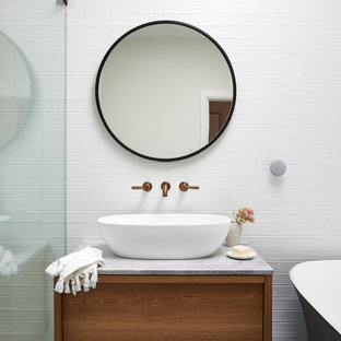 This is an example of a mid-sized beach style master bathroom in Sydney with furniture-like cabinets, medium wood cabinets, a freestanding tub, an open shower, a wall-mount toilet, white tile, subway tile, white walls, cement tiles, a vessel sink, marble benchtops, blue floor, a hinged shower door, grey benchtops, a laundry, a single vanity, a floating vanity, recessed and brick walls.