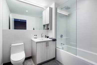 ニューヨークにある高級な中くらいなモダンスタイルのおしゃれなマスターバスルーム (フラットパネル扉のキャビネット、グレーのキャビネット、アルコーブ型浴槽、シャワー付き浴槽	、一体型トイレ	、グレーのタイル、セラミックタイル、白い壁、セラミックタイルの床、珪岩の洗面台、一体型シンク) の写真