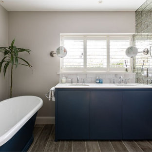 Medium sized contemporary ensuite bathroom in Buckinghamshire with flat-panel cabinets, blue cabinets, a freestanding bath, porcelain flooring, grey floors, white worktops, solid surface worktops, beige walls, a submerged sink, grey tiles, glass tiles, a wall niche, double sinks and a built in vanity unit.