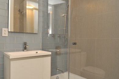 ニューヨークにある高級な中くらいなコンテンポラリースタイルのおしゃれなバスルーム (浴槽なし) (フラットパネル扉のキャビネット、白いキャビネット、アルコーブ型浴槽、シャワー付き浴槽	、一体型トイレ	、青いタイル、ガラスタイル、ベージュの壁、ラミネートの床、一体型シンク、人工大理石カウンター、ベージュの床、開き戸のシャワー) の写真