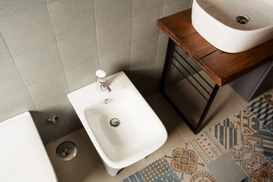 Bild på ett mellanstort funkis badrum, med skåp i slitet trä, ett platsbyggt badkar, en toalettstol med separat cisternkåpa, grå kakel, porslinskakel, grå väggar, klinkergolv i porslin, ett fristående handfat och träbänkskiva