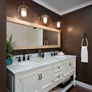 Modelo de cuarto de baño principal, costero, de tamaño medio, con lavabo bajoencimera, puertas de armario blancas, paredes marrones, sanitario de dos piezas, suelo de madera en tonos medios, encimera de mármol y armarios con paneles empotrados