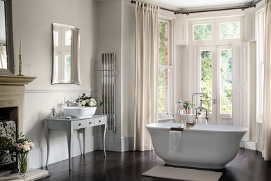 Inredning av ett modernt mellanstort en-suite badrum, med släta luckor, grå skåp, ett fristående badkar, grå väggar, mörkt trägolv, ett fristående handfat, bänkskiva i glas och brunt golv