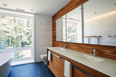 Inspiration for a medium sized modern ensuite bathroom in New York with flat-panel cabinets, medium wood cabinets, a freestanding bath, a built-in shower, a one-piece toilet, white tiles, stone tiles, white walls, porcelain flooring, an integrated sink, solid surface worktops, black floors, an open shower and white worktops.