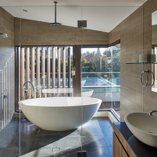 This is an example of a contemporary master wet room bathroom in Sydney with flat-panel cabinets, medium wood cabinets, a freestanding tub, brown tile, brown walls, a vessel sink, black floor, a hinged shower door and black benchtops.