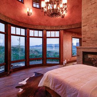Diseño de dormitorio principal, rústico, grande, con paredes rojas, suelo de madera en tonos medios, chimenea de doble cara, marco de chimenea de piedra y suelo marrón