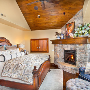 Imagen de dormitorio rural con paredes beige, suelo de madera en tonos medios, marco de chimenea de piedra y chimenea de esquina
