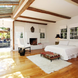 Modelo de dormitorio principal, mediterráneo, grande, con paredes blancas, chimenea tradicional, marco de chimenea de ladrillo y suelo de madera en tonos medios