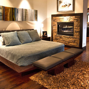 Ejemplo de dormitorio principal, minimalista, grande, con paredes beige, suelo de madera oscura, chimenea de doble cara y marco de chimenea de piedra