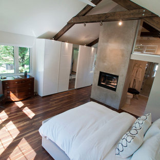 Foto de dormitorio principal, minimalista, de tamaño medio, con paredes blancas, suelo de madera en tonos medios, chimenea de doble cara, marco de chimenea de hormigón y suelo marrón