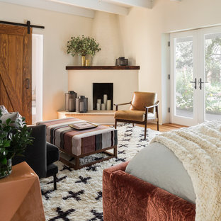 Diseño de dormitorio principal, moderno, con paredes blancas, suelo de madera en tonos medios, chimenea de esquina y marco de chimenea de ladrillo