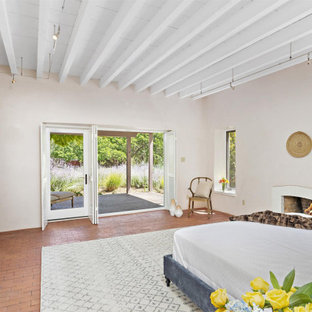 Modelo de dormitorio principal, de estilo americano, grande, con paredes beige, suelo de ladrillo, chimenea tradicional, marco de chimenea de ladrillo y suelo naranja