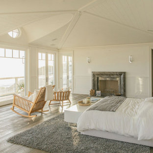 Geräumiges Maritimes Hauptschlafzimmer mit weißer Wandfarbe, hellem Holzboden, Kamin, Kaminumrandung aus Stein, grauem Boden, gewölbter Decke und vertäfelten Wänden in Los Angeles