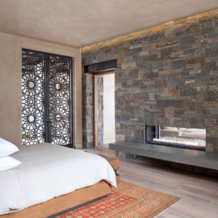 Diseño de dormitorio principal, actual, de tamaño medio, con paredes beige, suelo de madera clara, chimenea de doble cara, marco de chimenea de piedra y suelo beige