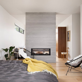 Modelo de dormitorio principal, actual, con paredes blancas, suelo de madera clara, marco de chimenea de hormigón, chimenea de doble cara y suelo beige