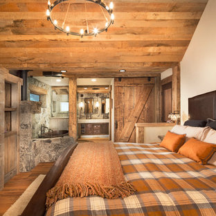 Modelo de dormitorio principal, rústico, de tamaño medio, con paredes beige, suelo de madera en tonos medios y marco de chimenea de piedra