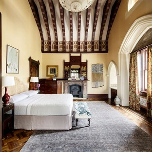 Modelo de dormitorio principal, tradicional, extra grande, con paredes amarillas, suelo de madera en tonos medios, estufa de leña, marco de chimenea de madera y suelo marrón