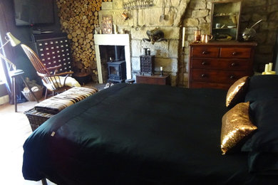 他の地域にある中くらいなエクレクティックスタイルのおしゃれな主寝室 (グレーの壁、無垢フローリング、薪ストーブ、石材の暖炉まわり、茶色い床)