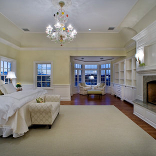 Großes Klassisches Hauptschlafzimmer mit gelber Wandfarbe, braunem Holzboden, Kamin, Kaminumrandung aus Stein und braunem Boden in New York