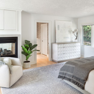 Modelo de dormitorio actual con paredes blancas, suelo de madera clara, chimenea de doble cara y suelo beige