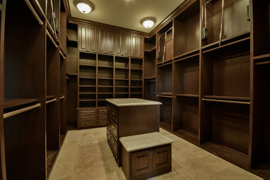This is an example of a large classic gender neutral walk-in wardrobe in Houston with raised-panel cabinets, brown cabinets, travertine flooring and beige floors.