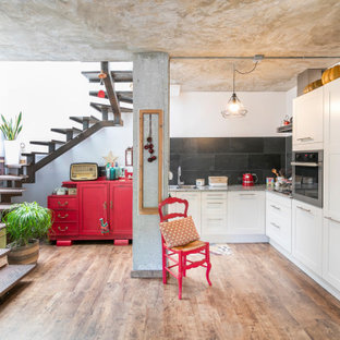 Imagen de cocina en L, ecléctica, sin isla, con armarios estilo shaker, puertas de armario blancas, salpicadero negro, electrodomésticos blancos, suelo de madera en tonos medios, suelo marrón y encimeras grises
