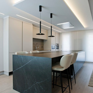 Foto de cocina lineal, actual, grande, abierta, con armarios con paneles lisos, electrodomésticos de acero inoxidable, una isla, puertas de armario blancas, salpicadero verde, salpicadero de vidrio templado, suelo de madera clara, suelo beige y encimeras negras