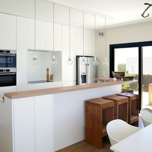 Modelo de cocina comedor minimalista con electrodomésticos de acero inoxidable, suelo de madera en tonos medios, encimeras blancas, una isla, armarios con paneles lisos, puertas de armario blancas y salpicadero blanco