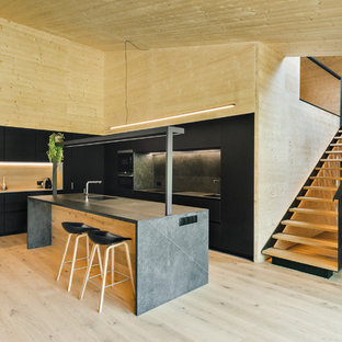 Ejemplo de cocina en L, minimalista, abierta, con fregadero integrado, armarios con paneles lisos, puertas de armario negras, salpicadero negro, electrodomésticos negros, suelo de madera clara, una isla y encimeras negras