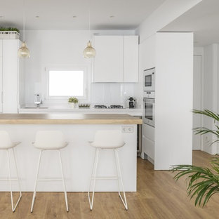 Ejemplo de cocina en L, contemporánea, con armarios con paneles lisos, puertas de armario blancas, encimera de madera, salpicadero blanco, electrodomésticos blancos, una isla, fregadero bajoencimera, salpicadero de vidrio templado, suelo de madera clara, suelo marrón y encimeras beige
