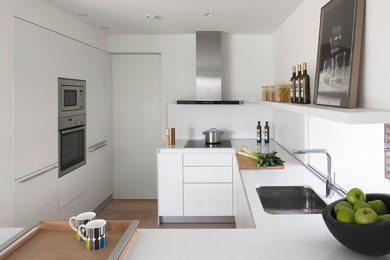 Idéer för ett litet modernt kök, med en enkel diskho, släta luckor, vita skåp, vitt stänkskydd, rostfria vitvaror, mellanmörkt trägolv och en halv köksö