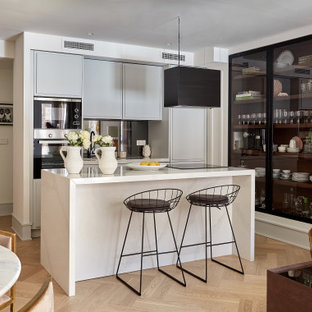 Foto de cocina de galera, actual, pequeña, abierta, con armarios con paneles lisos, puertas de armario grises, electrodomésticos de acero inoxidable, suelo de madera clara, una isla, suelo beige y encimeras blancas
