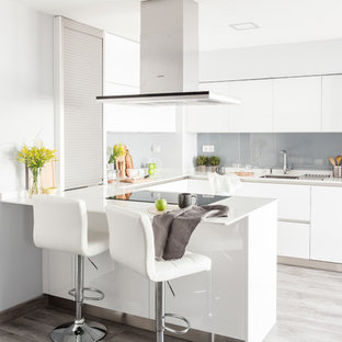 Modelo de cocina en U, contemporánea, de tamaño medio, con fregadero bajoencimera, armarios con paneles lisos, puertas de armario blancas, encimera de laminado, salpicadero verde, electrodomésticos de acero inoxidable, suelo laminado, península, suelo gris, encimeras blancas y salpicadero de vidrio templado