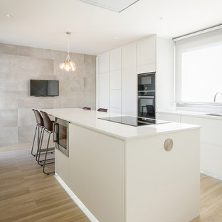 Ejemplo de cocina de galera, contemporánea, con armarios con paneles lisos, puertas de armario blancas, encimera de cuarzo compacto, electrodomésticos negros, una isla, encimeras blancas, suelo de madera clara y suelo beige