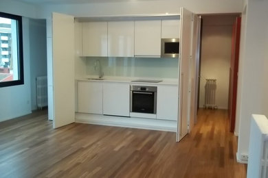 Foto de cocina lineal minimalista de tamaño medio abierta sin isla con fregadero bajoencimera, armarios con paneles lisos, puertas de armario blancas, salpicadero blanco, electrodomésticos de acero inoxidable, suelo de madera en tonos medios y salpicadero de vidrio templado