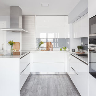 Modelo de cocina en U, actual, con fregadero bajoencimera, armarios con paneles lisos, puertas de armario blancas, salpicadero verde, salpicadero de vidrio templado, electrodomésticos negros, península, suelo gris y encimeras blancas